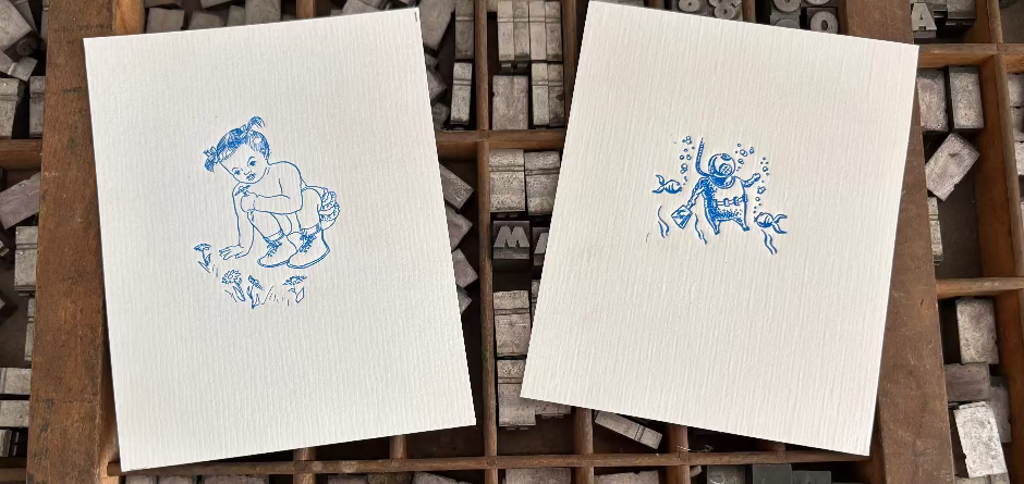 Two letterpress cards with blue illustrations on a wooden printing block tray.